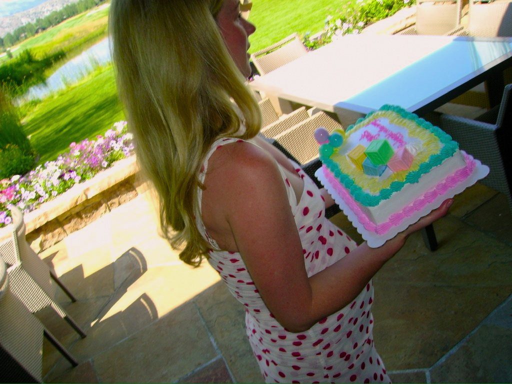 From Kara Hobby’s Wilson Mom summer: woman carrying a pastel baby-shower cake on a patio