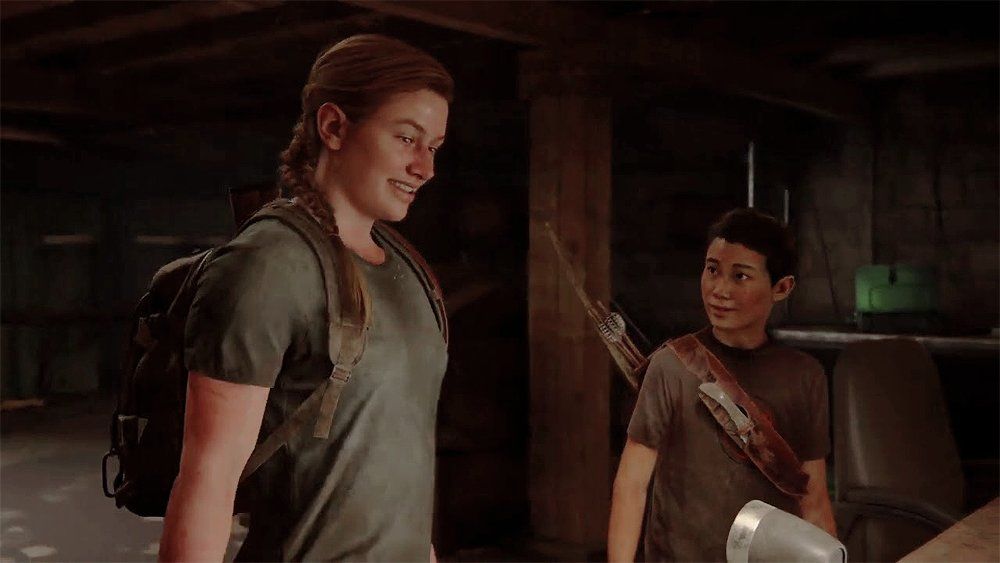 Abby (left) and Lev (right) in a scene from the video game 'The Last of Us, Part II' (2020). Abby is a white teenage girl with a muscular build and her blonde hair pulled back into a braid. She's wearing a grey t-shirt and a brown backpack. She's smiling as she looks down at something. Lev is an Asian trans boy who is shorter than Abby. He has close-cropped black hair and is wearing a brownish t-shirt with a quiver of arrows and a bow strapped to his back. He's looking at Abby. They are in a basement standing at a table with a radio on it.
