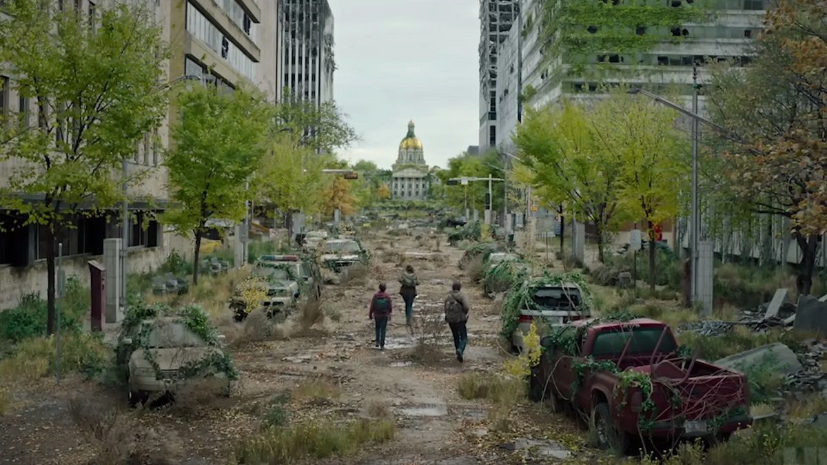An image of post-apocalyptic downtown Boston. The gold-domed State House is in the distance at the end of a long street that is covered in overgrown foliage that half buries wrecked, abandoned cars. We see the small figures of Ellie, Joel, and Tess walking toward the State House.