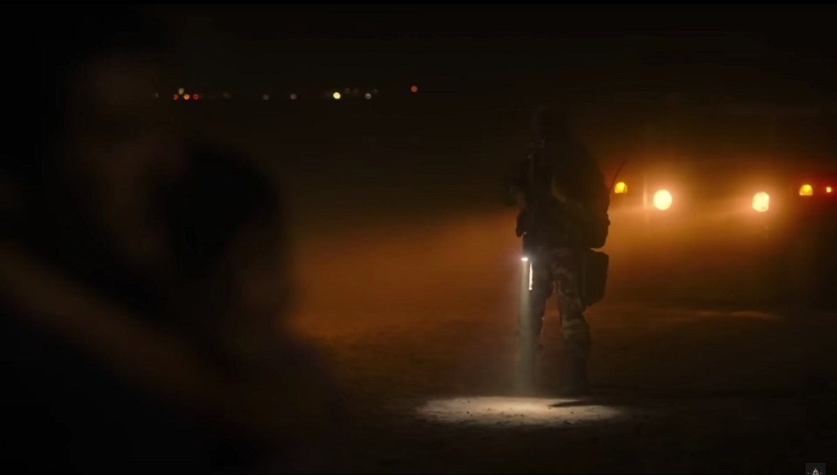 A dark, evening image from the last of us pilot. In the foreground, we see the shadowy silhouette of Joel and Sarah. In the background, in focus, we see a masked, helmeted soldier standing in the headlights of a truck. He's holding a gun and a flashlight, which creates a pool of light at his feet.