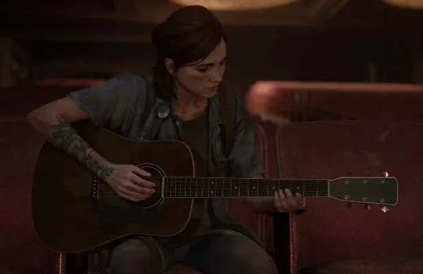 Ellie plays guitar in a Seattle theater in 'The Last of Us, Part II' (2020). Ellie is sitting in one of the red seats in an empty theater playing guitar. She is a young white woman with chin-length brown hair, and wears a short-sleeved blue button down over a grey t-shirt and jeans. She has a large tattoo on her right arm.