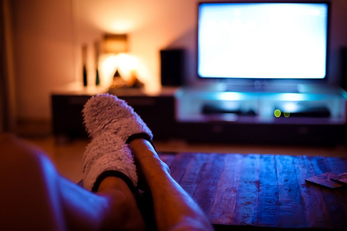Stock image of someone with their slippered feet up on a wooden coffee table watching TV.