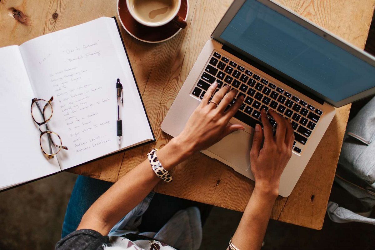 Stock image of a woman typing on a laptop. The shot is from overhead, so we only see her arms. She's seated at a wooden table, and there's an open notebook sitting to her left with a pen and her glasses on it, and a cup of coffee just above that.