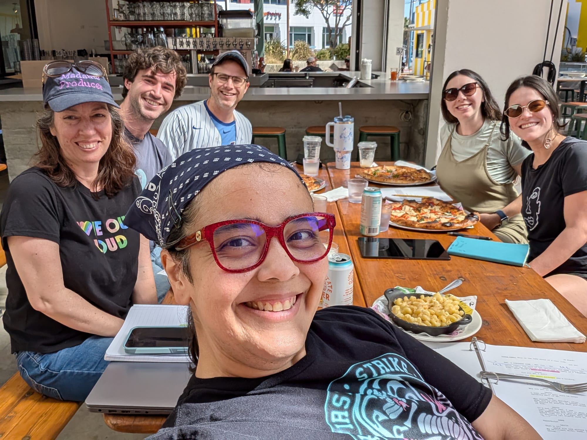 Photo of six people seated around a long, wooden picnic table at an outdoor restaurant smiling into the camera