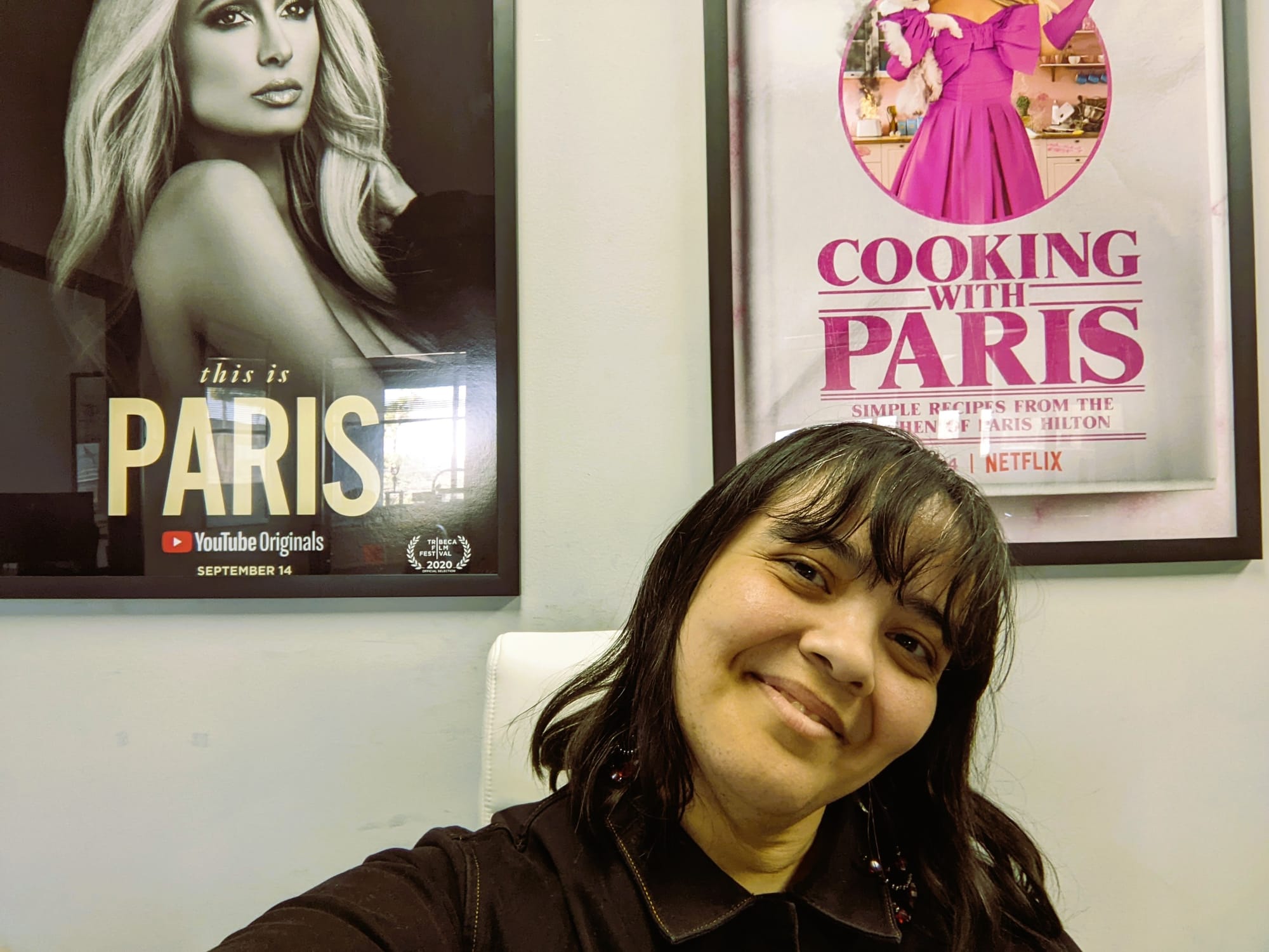 Image of Teresa Jusino (Latina with chin-length dark hair and bangs) wearing a black jumpsuit and sitting in a white chair with two Paris Hilton show-related posters in frames on the wall behind her.