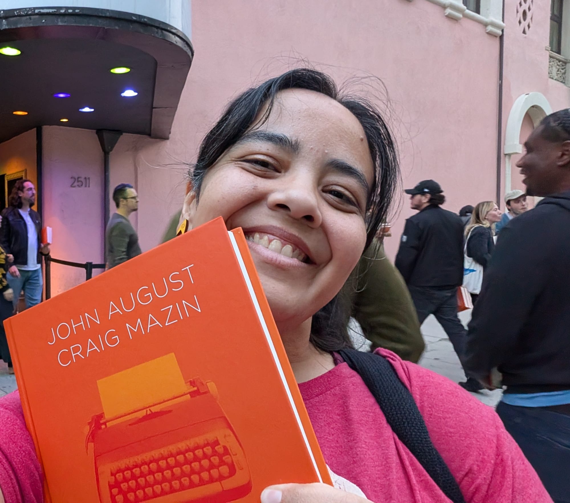 Image of Teresa Jusino (a Latina with dark hair) wearing a red t-shirt smiling as she holds up a copy of the 'Scriptnotes' book.