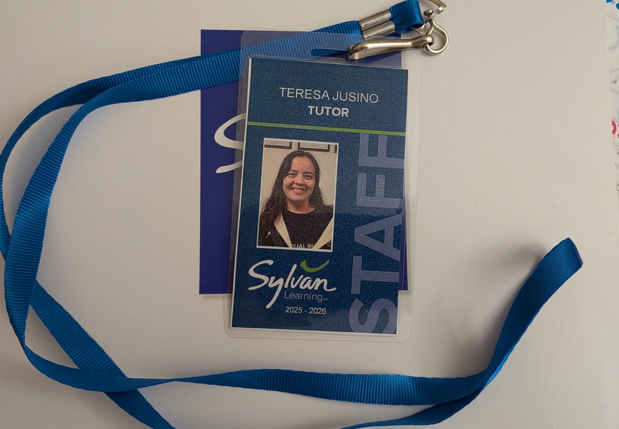 Image of a blue photo ID badge on a blue lanyard sitting on a white folder. The badge reads "Teresa Jusino - Tutor - Sylvan Learning 2025-2026"
