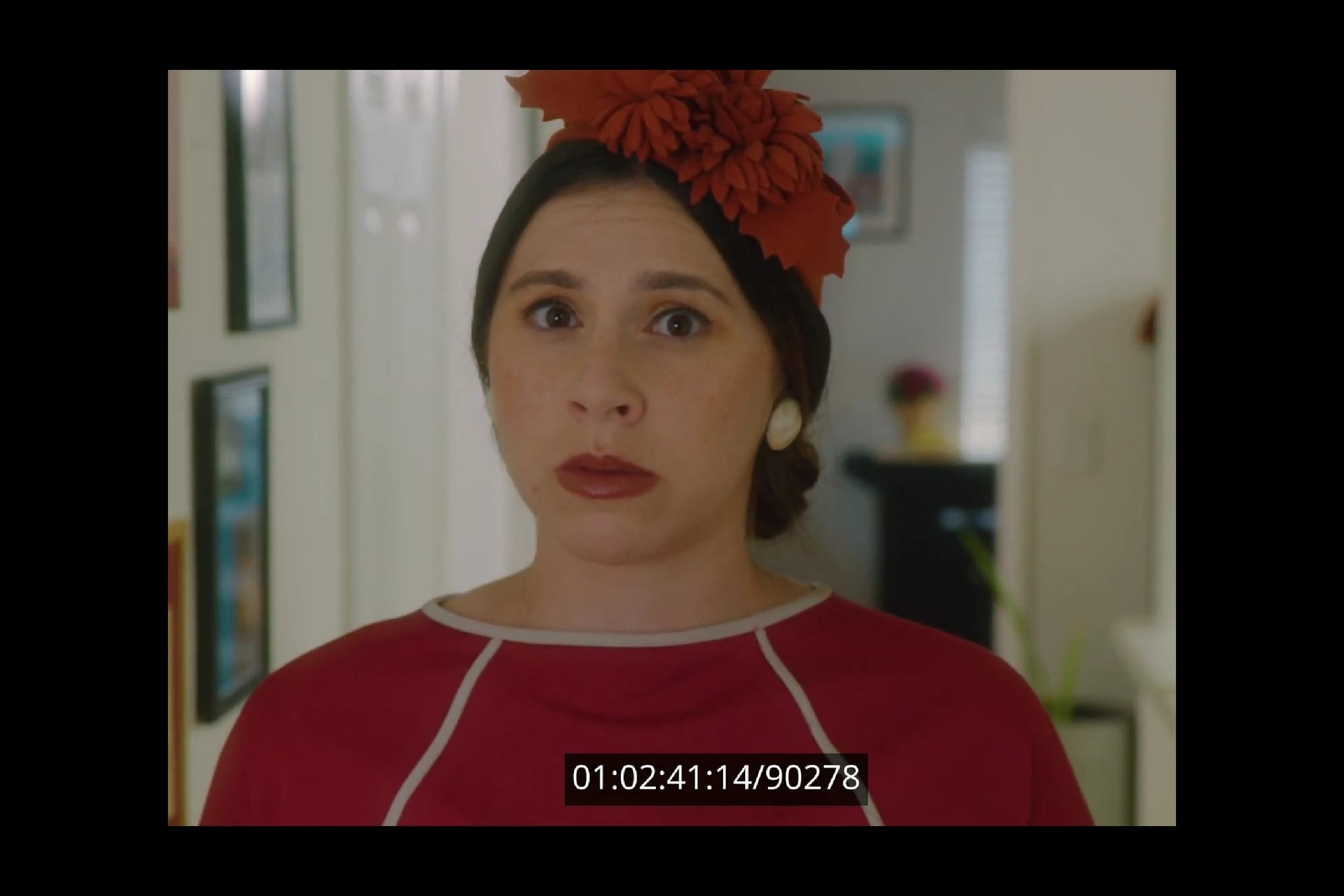 Image of a time-stamped still from a short film featuring a close-up of a white woman with dark hair pulled back wearing a 1940s flowered hat and a red dress. She looks worried.