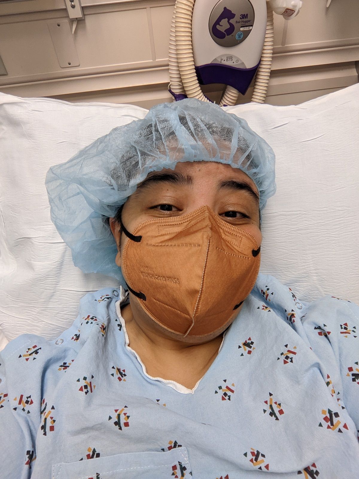 Teresa lying on a white pillow in a hospital bed in a blue hair cap, brown KN95 mask, and a blue hospital gown.
