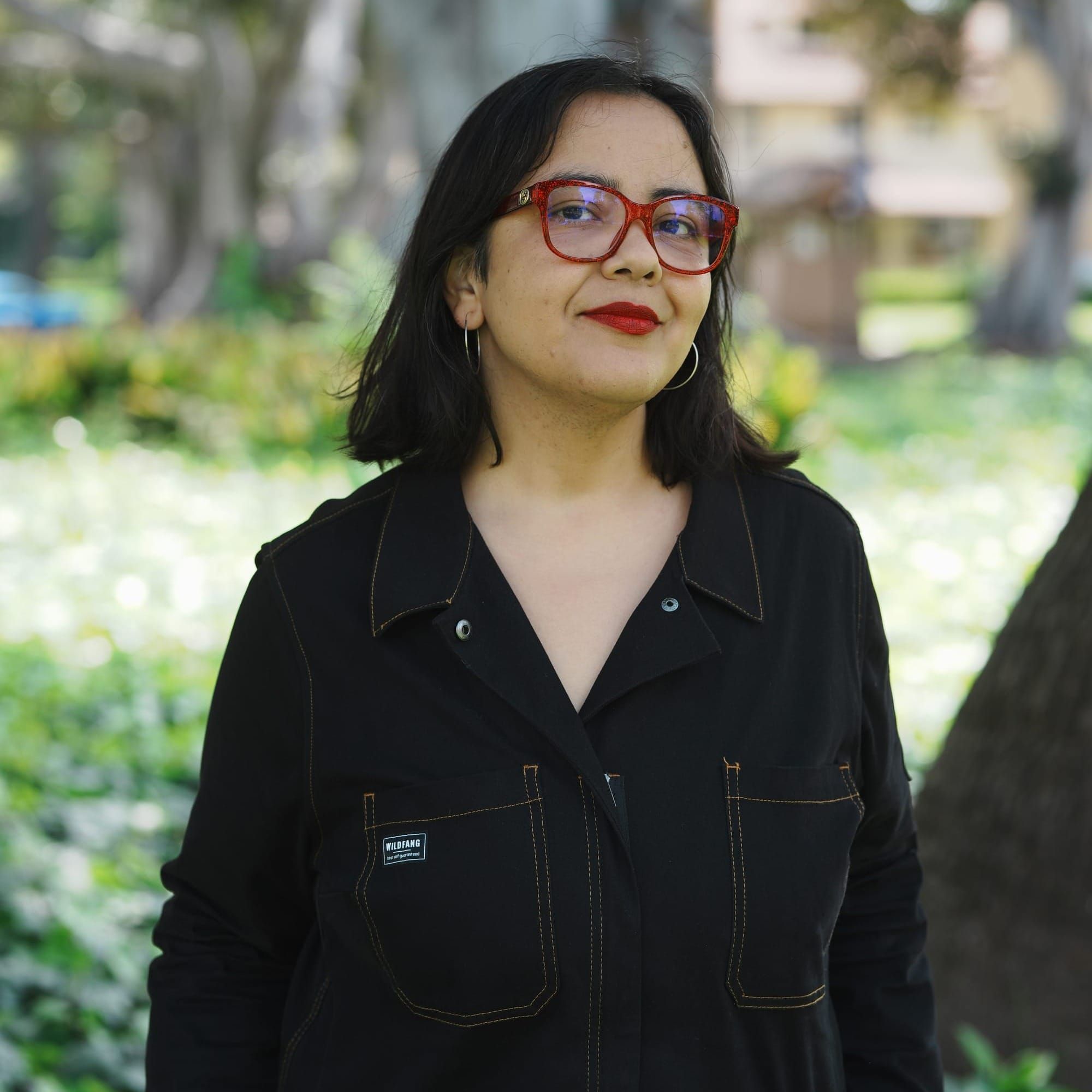 A Latina w/shoulder-length dark hair, glasses with red frames, silver hoop earrings, and a black jumpsuit. 
