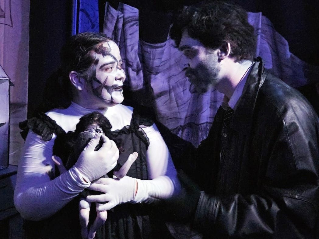 A woman holding a doll & a man looking at each other, both wearing black & white clothes & make-up.