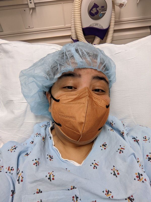 Teresa lying on a white pillow in a hospital bed in a blue hair cap, brown KN95 mask, and a blue hospital gown.