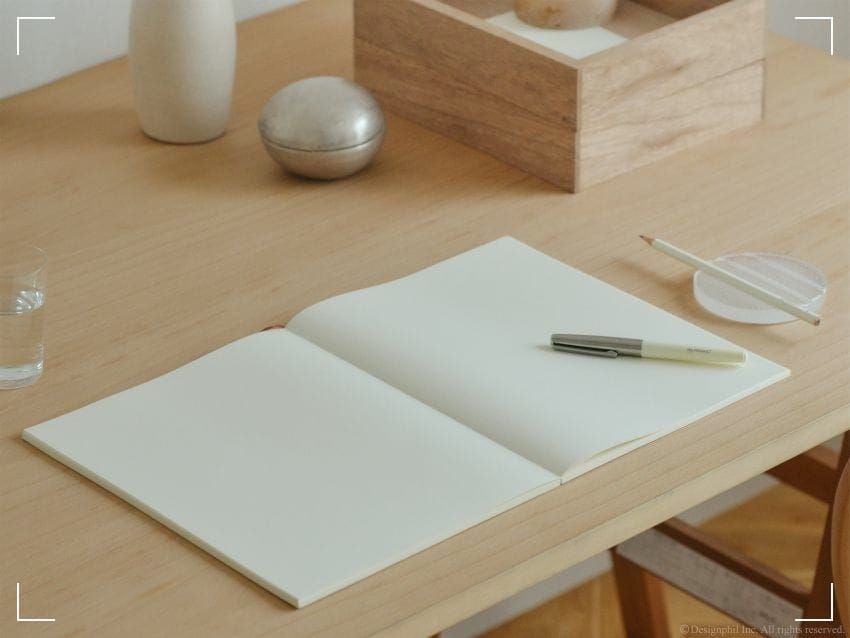 An open Midori MD paper notebook lying open and flat on a desk with some writing accessories included.