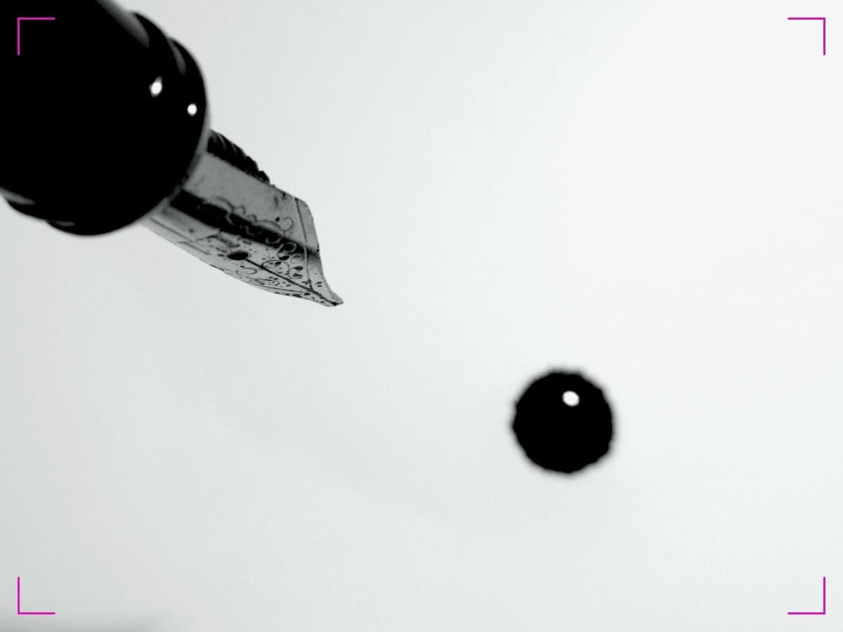 A fountain pen with the nib in close-up and a drop of ink on the white paper.