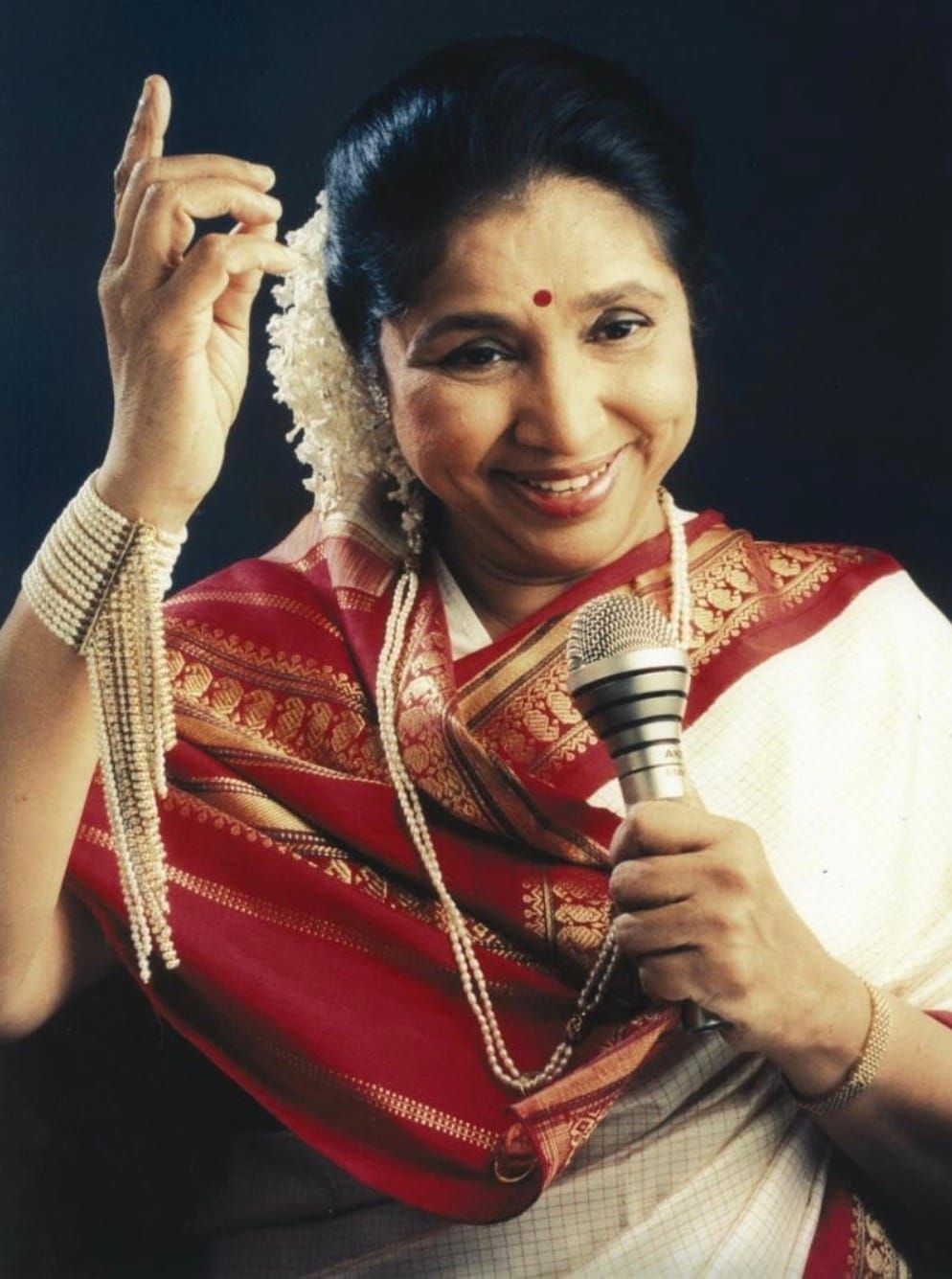 Asha Bhosle smiling, holding a microphone, in a red and gold sari.