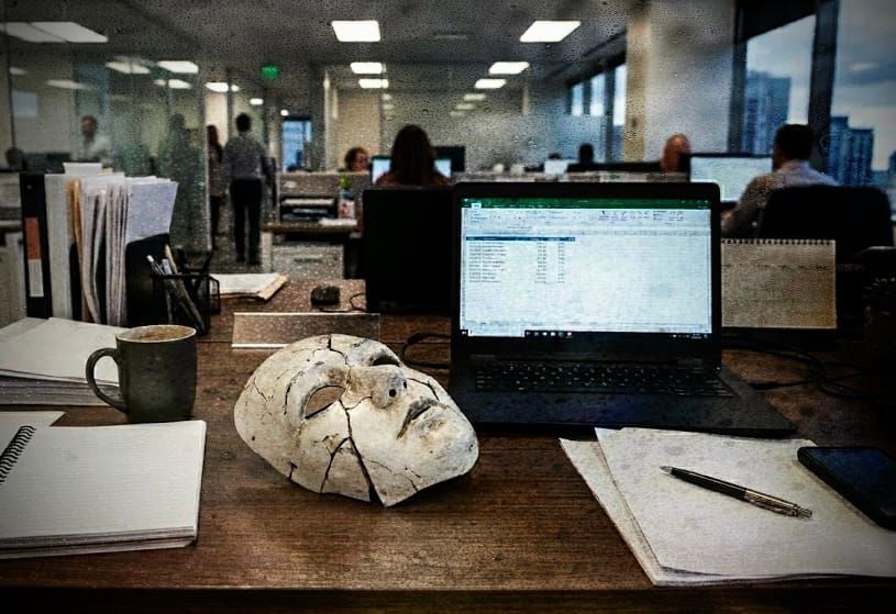 A cracked plaster face mask rests on a wooden office desk, surrounded by an open laptop displaying a spreadsheet, a coffee mug, notebooks, and a pen.