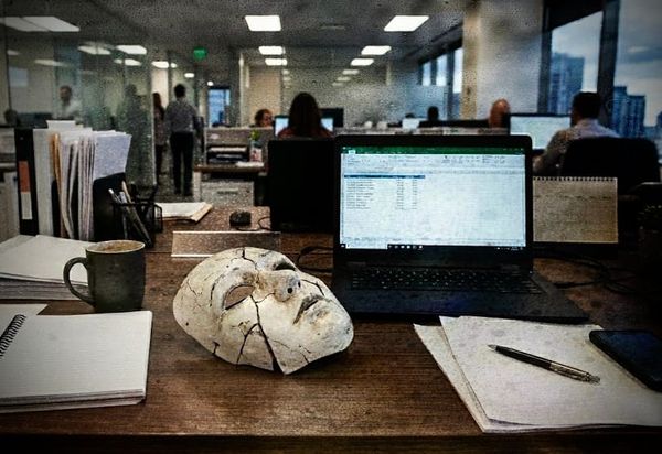 A cracked plaster face mask rests on a wooden office desk, surrounded by an open laptop displaying a spreadsheet, a coffee mug, notebooks, and a pen.