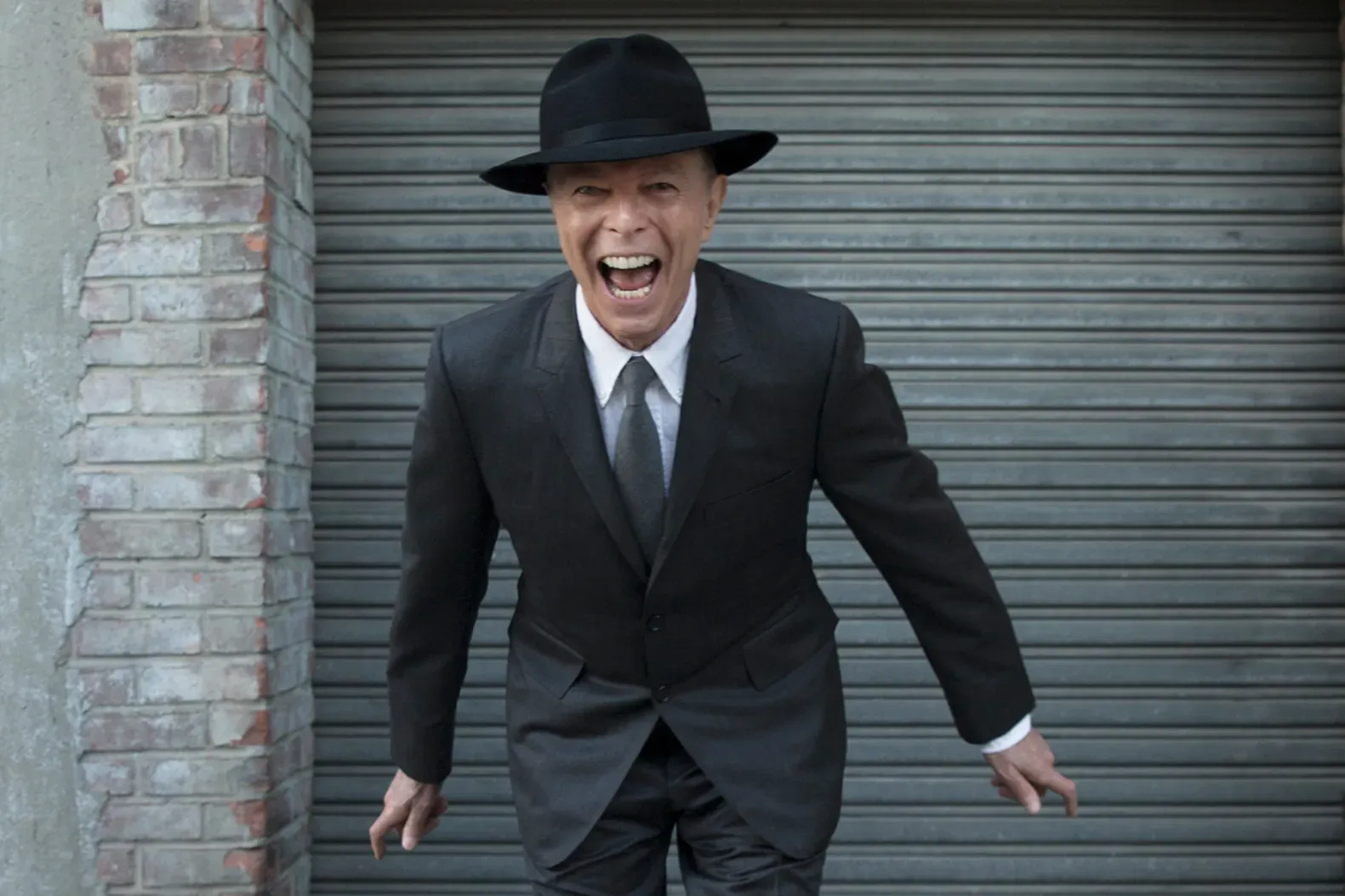 David Bowie leans forward mid-shout, wearing a black suit, white shirt, dark tie and fedora, before a grey metal shutter and brick wall, promoting Blackstar.