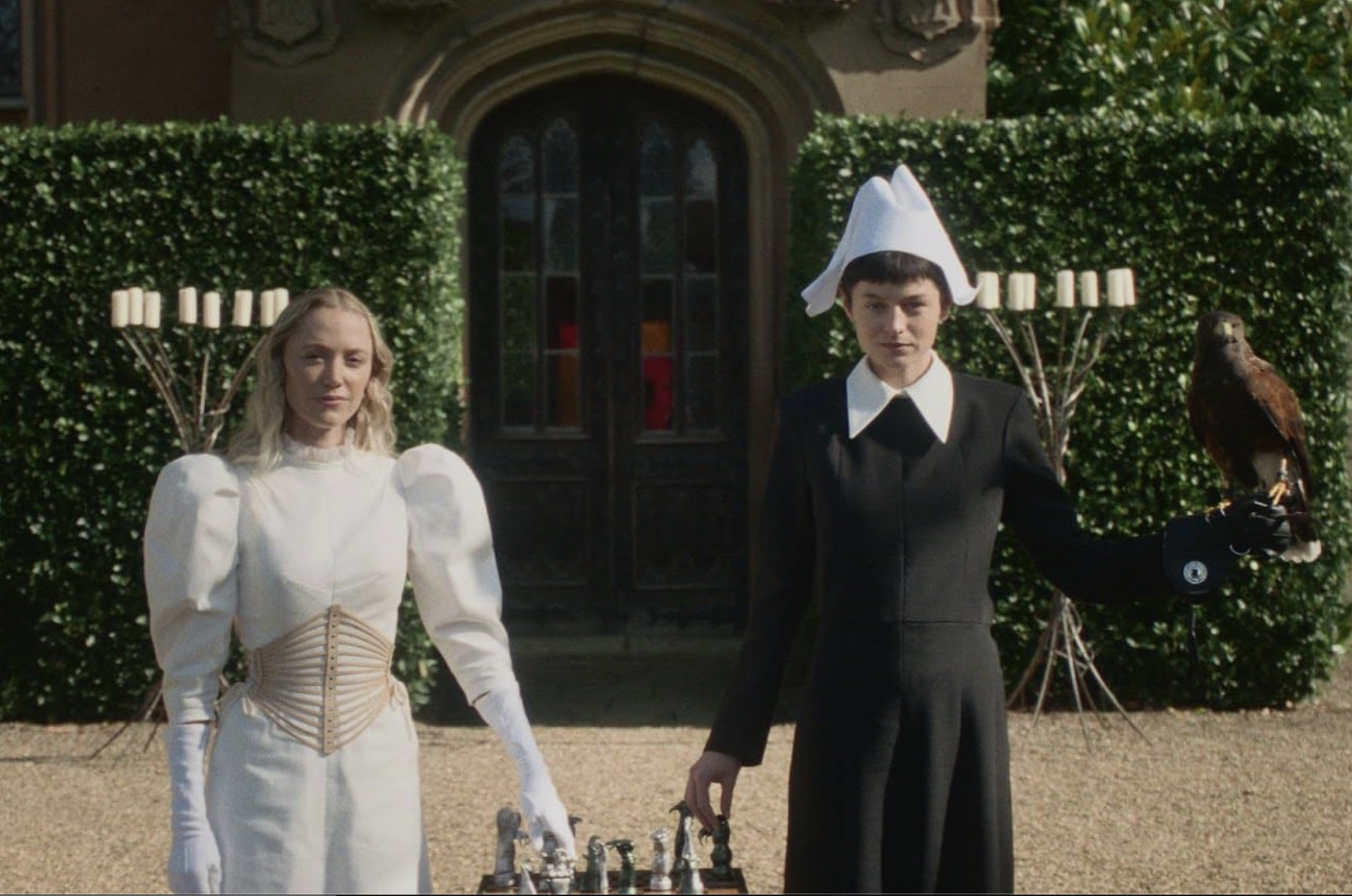 Two women stand in front of a mansion. One holds a falcon, and both touch pieces on a chess board.