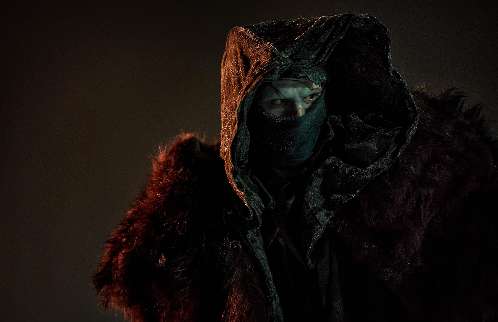 A man is shrouded in a cowl and deep shadows. 