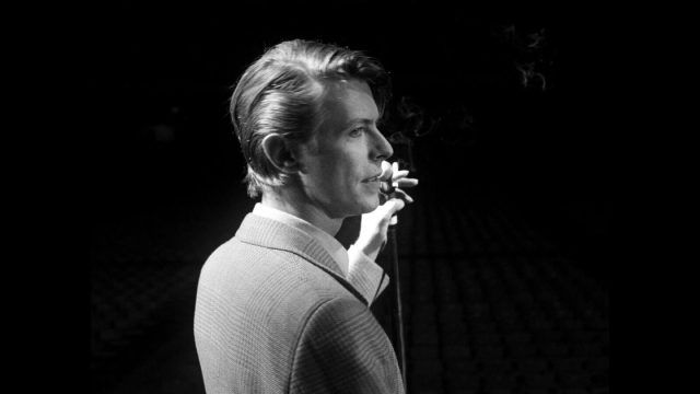 David Bowie, turned away from the camera, holds a microphone in a black-and-white still from MOONAGE DAYDREAM.