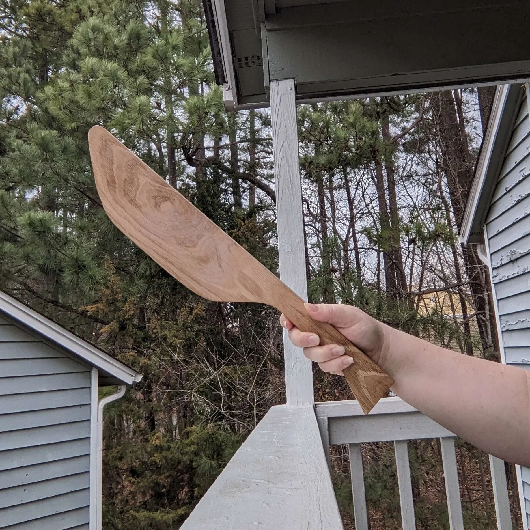 A hand is holding a wooden paddle shaped like a giant curved knife.