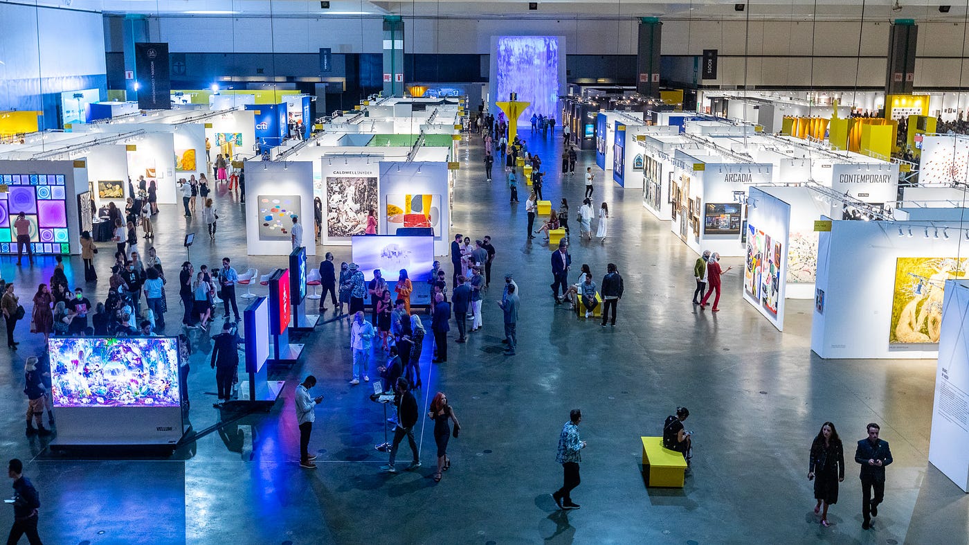 Lanes of white booths create a grid in a large convention center. Various colorful artworks hang on the walls. A row of benches fills the widest central pathway between booths. Observers walk about.