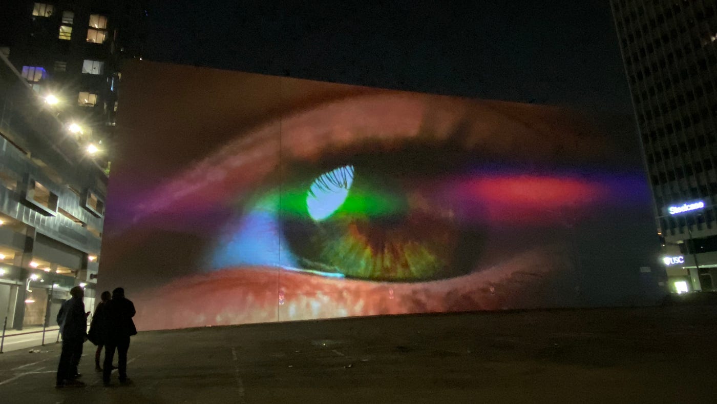 A moment from Sarah Rara’s Screen Lovers: An eye projected at about four times the height of a person on a wall outside at night.