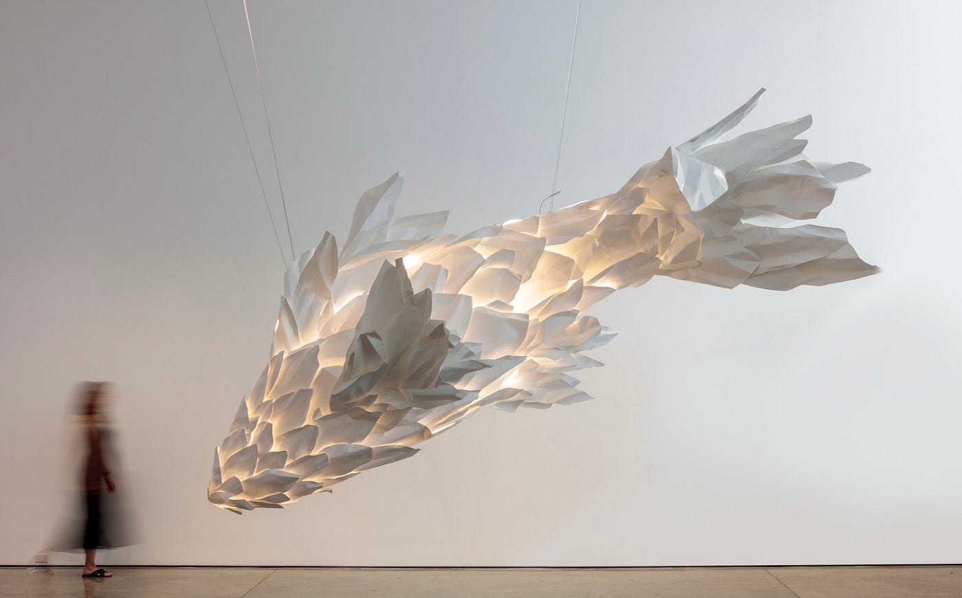 Against the backdrop of a white gallery wall, an enormous sculpture is suspended in air. The sculpture is also white and its assembly creates the shape of a fish, like a koi or betta fish with its draping, flared fins. The thin spaces in between the fish’s scales are illuminated from within. Off to the side a woman, blurred and in-motion, is a fraction of the size of the sculpture.