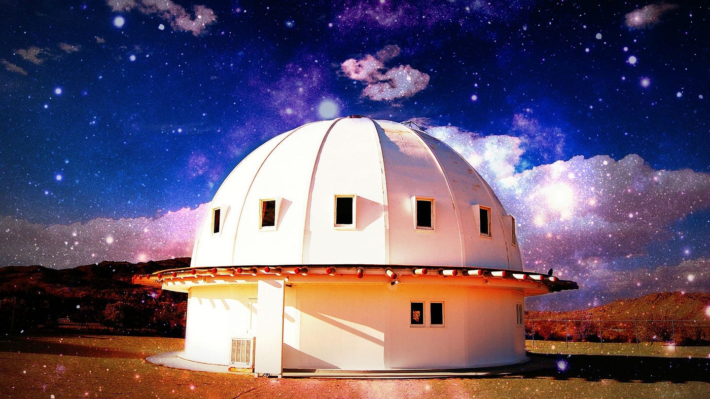 There’s a large structure in the image center, set in a desert landscape. It’s completely white and domed. There are symmetrical sections of the dome, like slices of cake, and in each one is a small window. The sky is both bright and night-like. A filter has been layered into the image so that it has a dreamlike quality; it looks as though stars have been superimposed on the full photo.