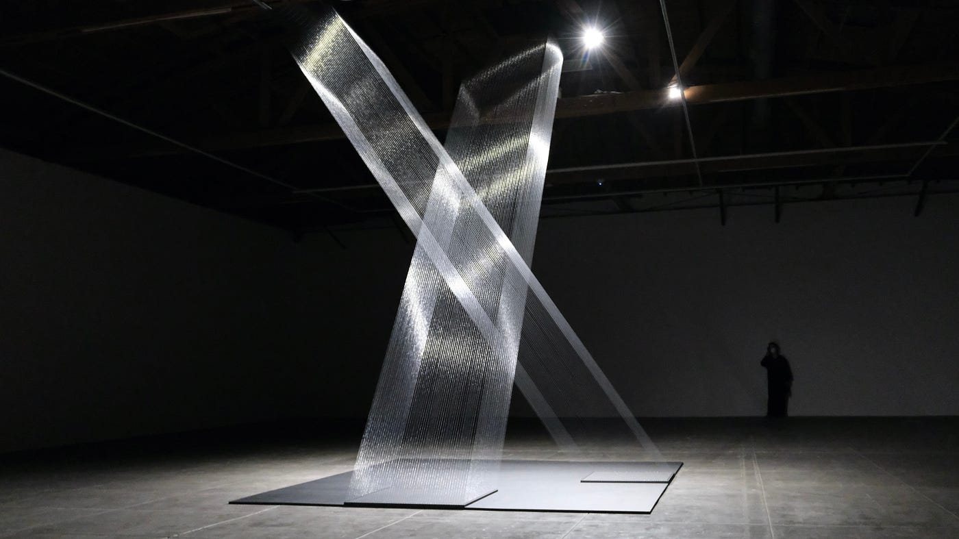 The gallery is a large, darkened space, more like a warehouse than a traditional gallery. In its center is a massive, delicate sculpture made of silver thread, light by a few overhead spotlights. There are two columns of this thread and then look more like woven steel than thread. The columns intersect and their intersection creates shapes that morph as the viewer’s angle shifts; sometimes the overlap creates a diamond or a tilted rectangle. A person hovers in shadow along the back wall.