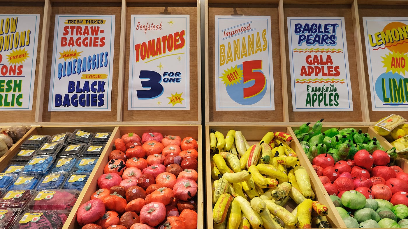 A typical supermarket vista — or is it? Wood crating holds ordinary produce: bananas, berries, apples, and tomatoes. Signage above includes pricing and declaratives such as “Fresh!” But the signs also include text like “baglet pears” and “bluebaggies.” Upon closer inspection, the produce is entirely composed of colorful, single-use plastic bags.