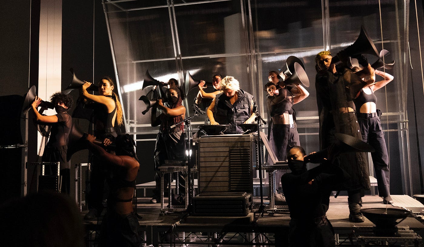 Performers dressed in all-black face front, standing on a stage. The setting is stark and industrial: concrete columns flank the dimly lit view and the bare-bones stage makes no effort to conceal the scaffolding underneath. There’s a textured backdrop of ribbed plexiglass. The performers hold black megaphones up to their ears.