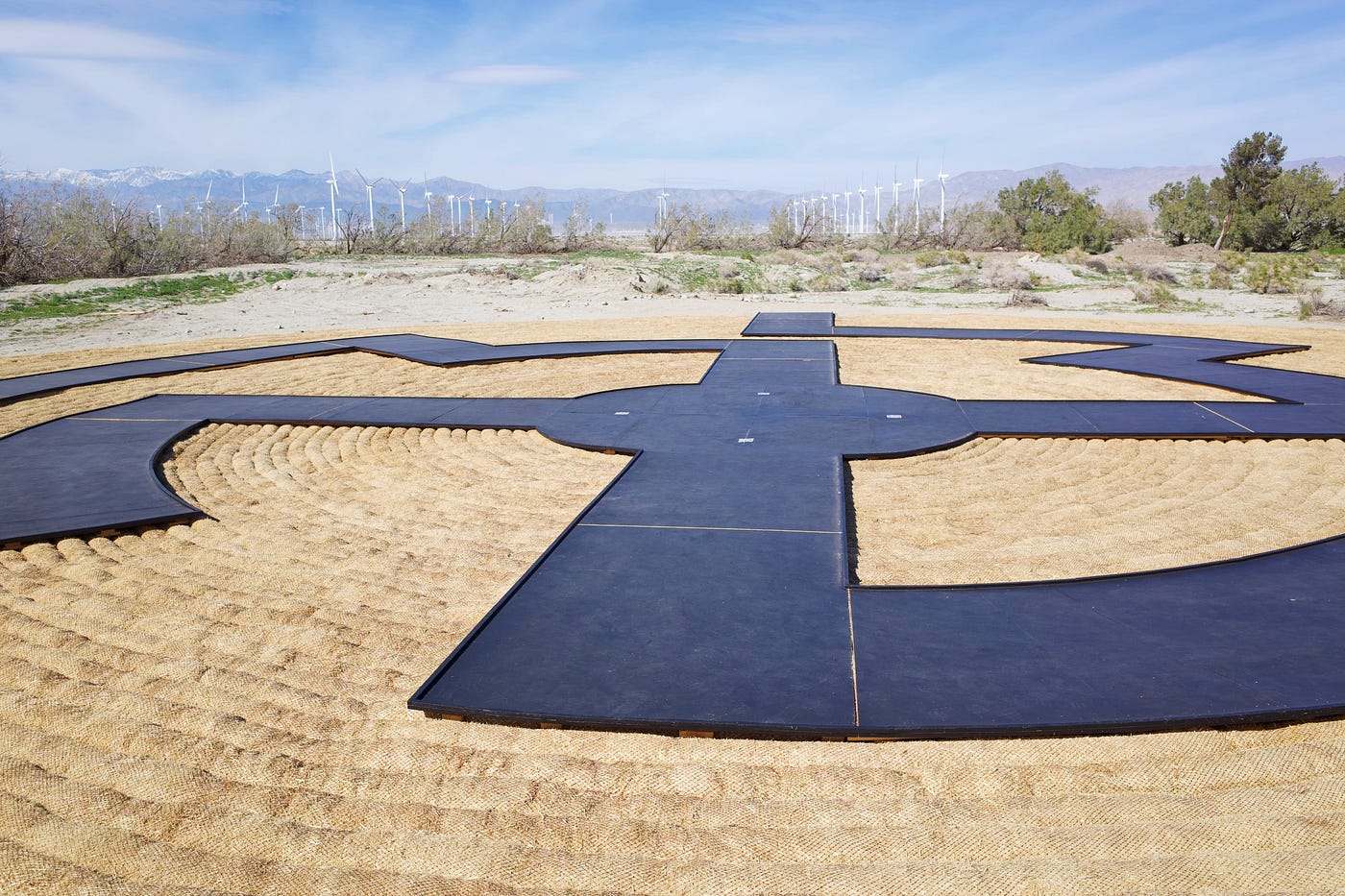 Rows of wind turbines are of in the distance. In the foreground is a large, woven sculpture. Coils of basket-weaving material form a flat disc. On top of this is a black, maze-like pathway.