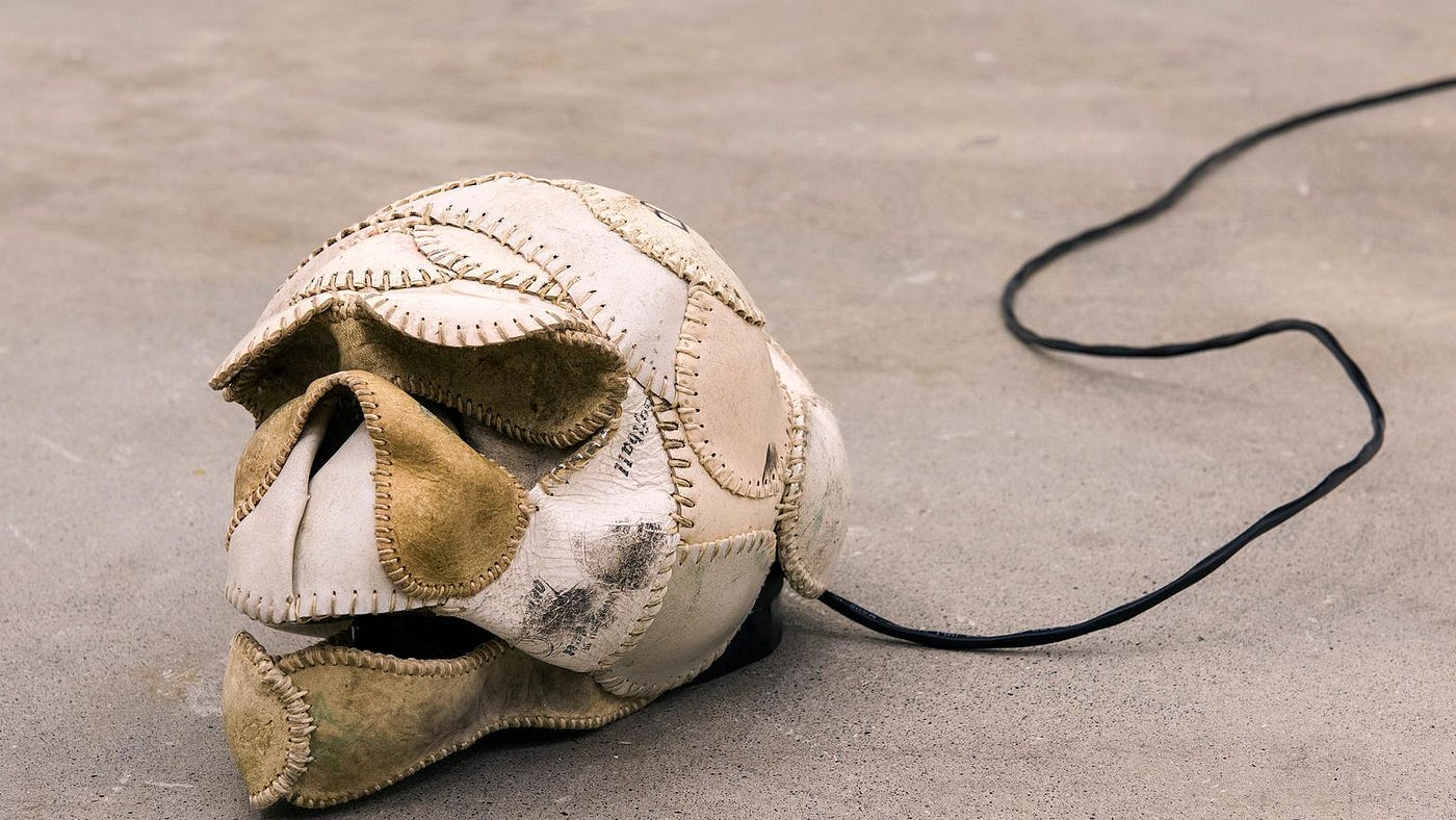 A close-up of leather pieces taken from baseballs or softballs and now stitched together. Some of the pieces are white and smooth; some are darker, grey or tan, more aged and worn. The stitching is sometimes narrow and delicate, and other times gaping. These leather pieces form the shape of a skull.
