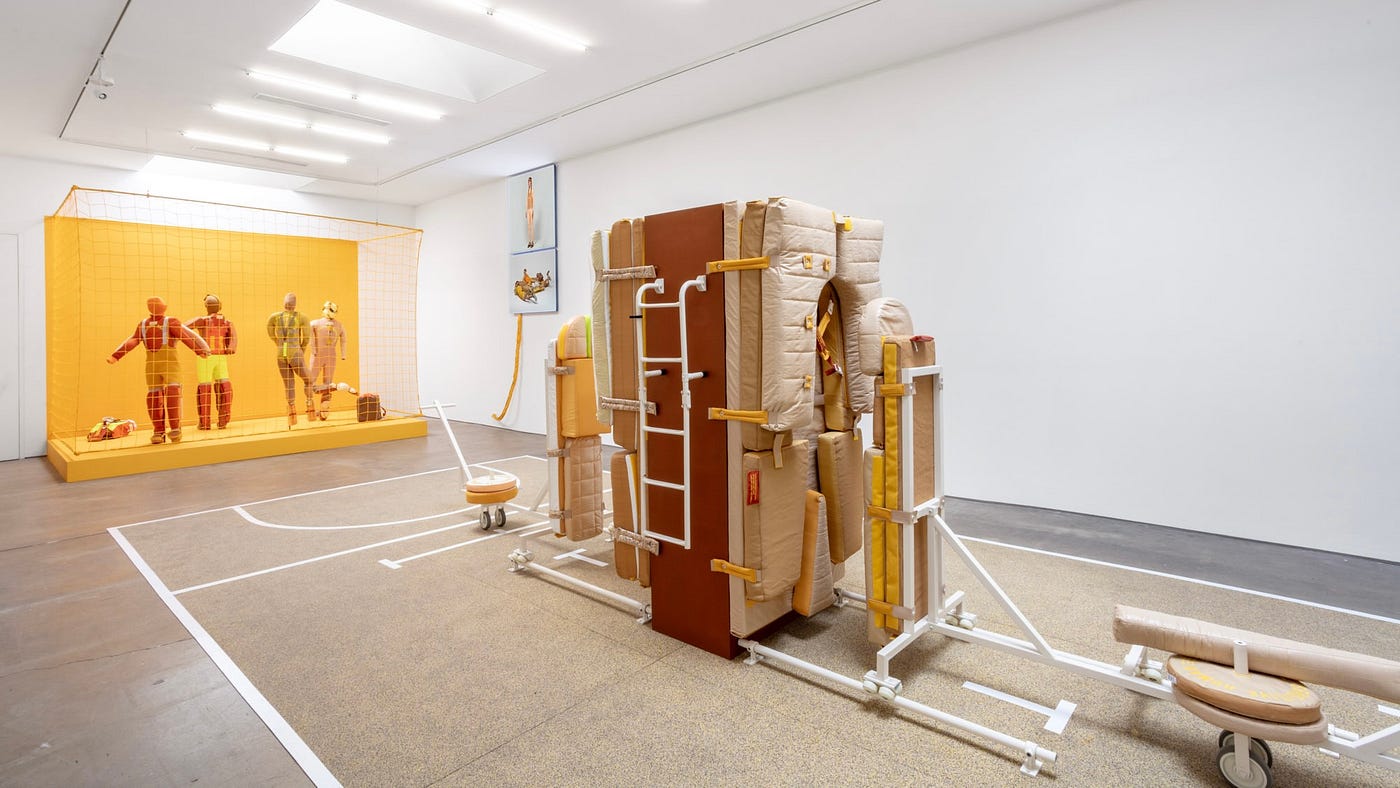 A white-walled gallery space. At the far end of the room is a bright yellow platform, surrounded by yellow netting. Inside are four mannequins wearing orange and yellow clothing that looks like a mix of sports uniforms and hi-vis work wear. In the foreground is a large contraption on a modified basketball court, resembling a combination of weight lifting bench and a vertically-oriented sleeper sofa.