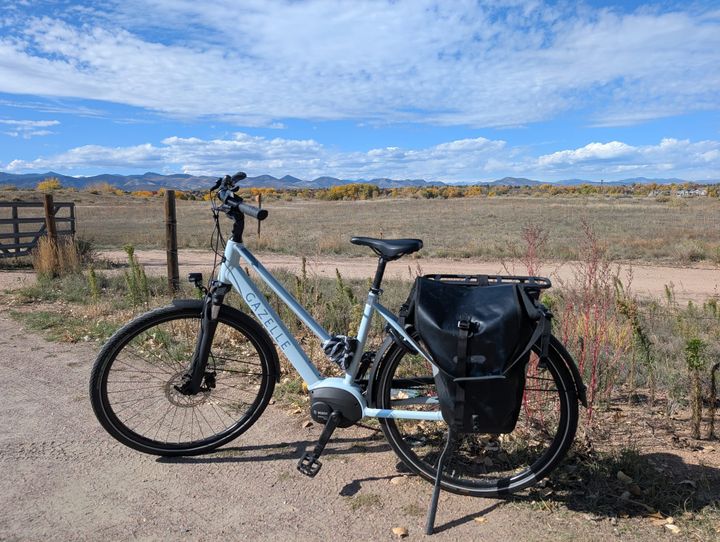 My blue gazelle medeo t9 ebike with an open Ortlieb pannier on the back, resting on its kickstand on a packed light gravel trail in front of a field with mountains in the distance