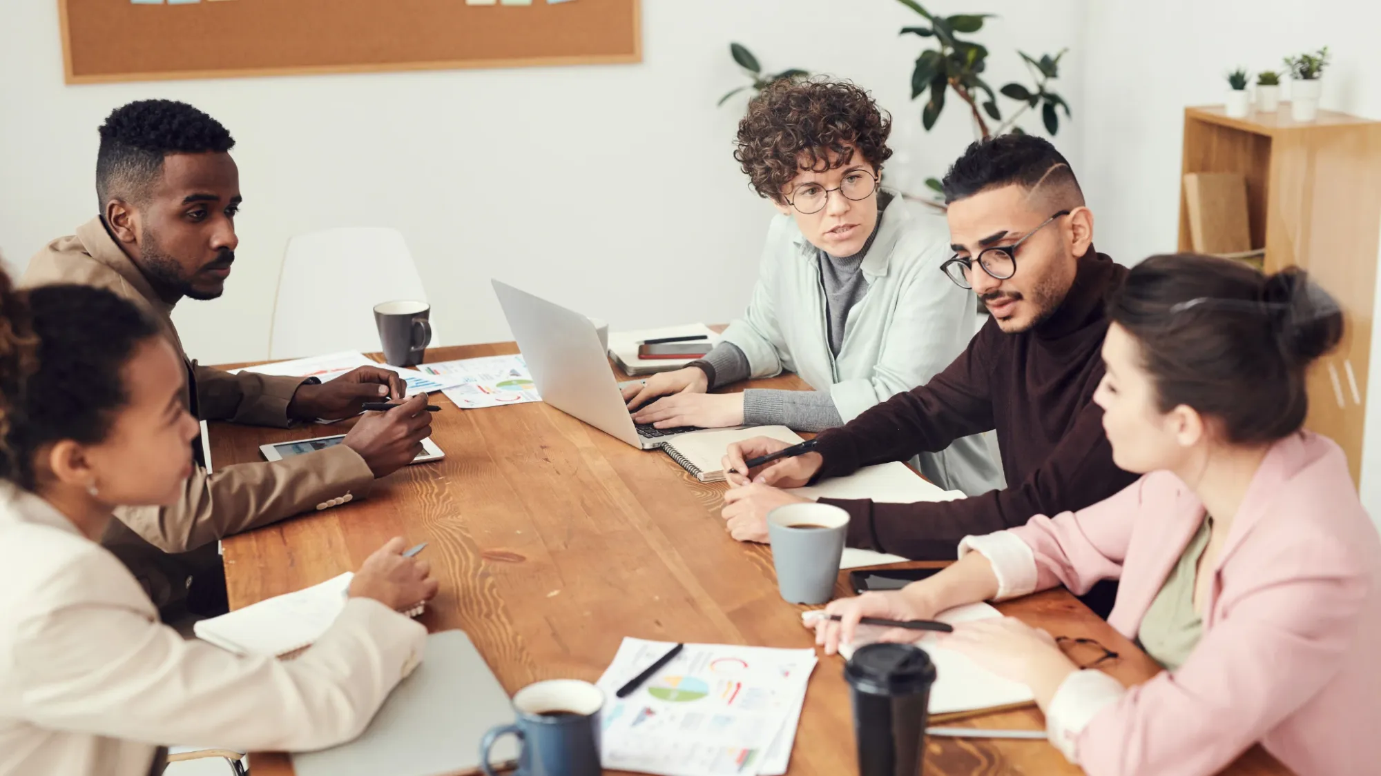 A diverse group of people sitting around a large wooden table in an office, engaged in a meeting or discussion. They are looking at laptops and documents, suggesting collaboration.
