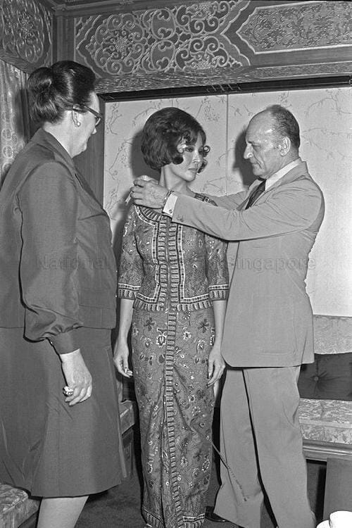 Black and white photo of fashion designer Pierre Balmain fitting a Singapore Airlines stewardess uniform on a young woman, as another woman observes