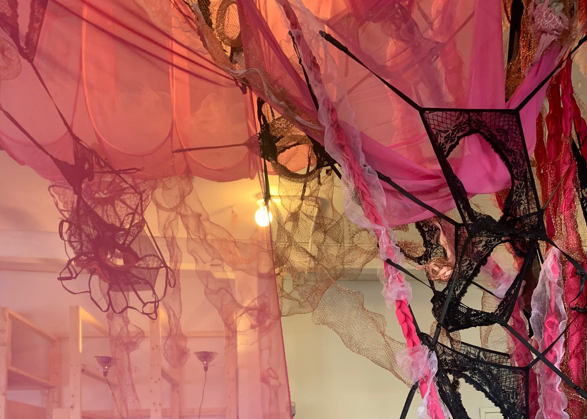 A shrine-like installation made from pink netting, black lace underwear, and suspended domestic textiles. No figure is present. The structure resembles an erotic web, layered and ceremonial.