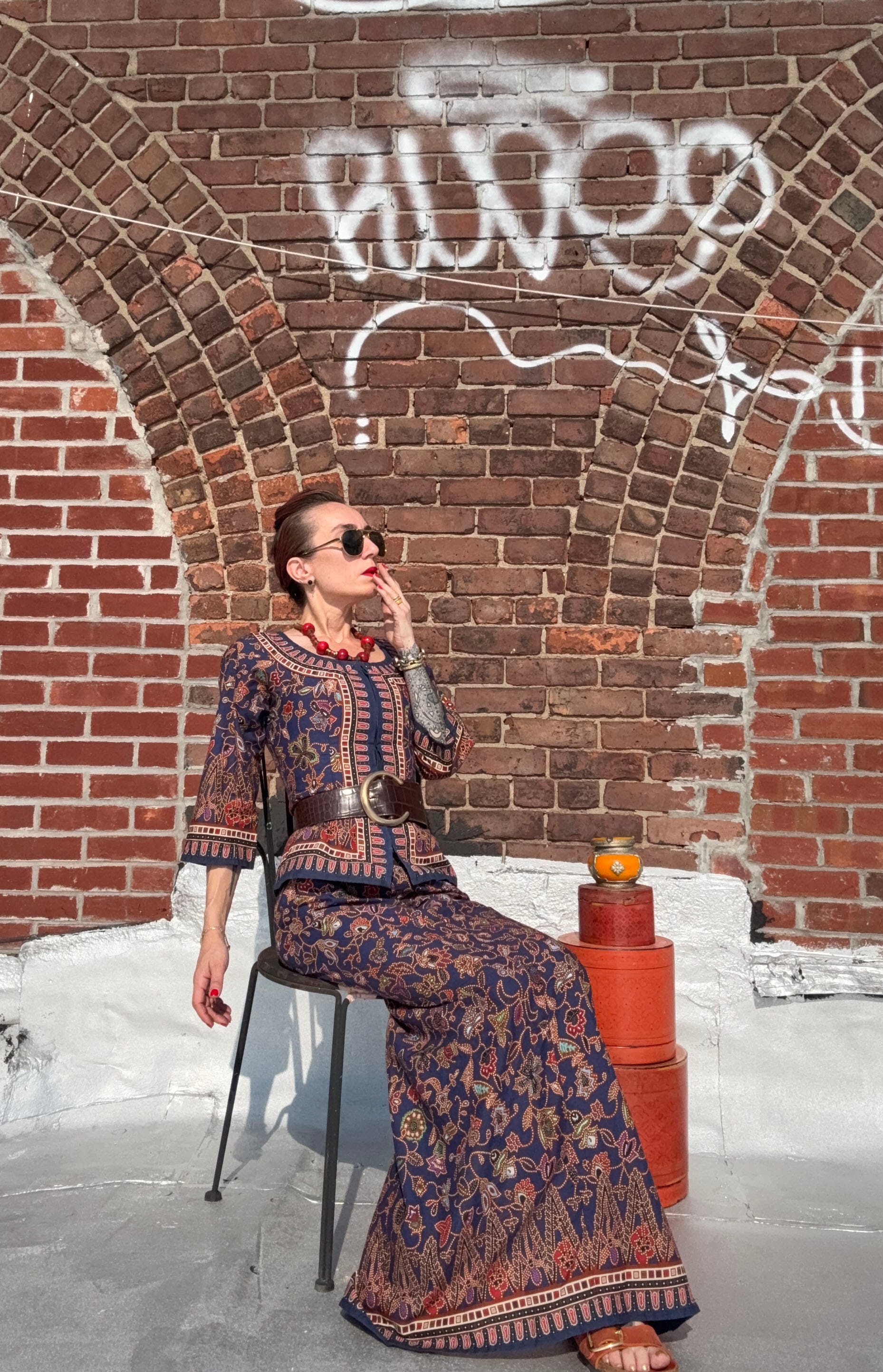 The artist sits against a graffitied brick wall on a Brooklyn rooftop, wearing a vintage Singapore Airlines sarong kebaya set in blue and red, styled with a wide belt, red jewelry, and sandals. A lacquered red tiffin and teacup rest beside her. Sunlight and shadow animate the quiet composition.