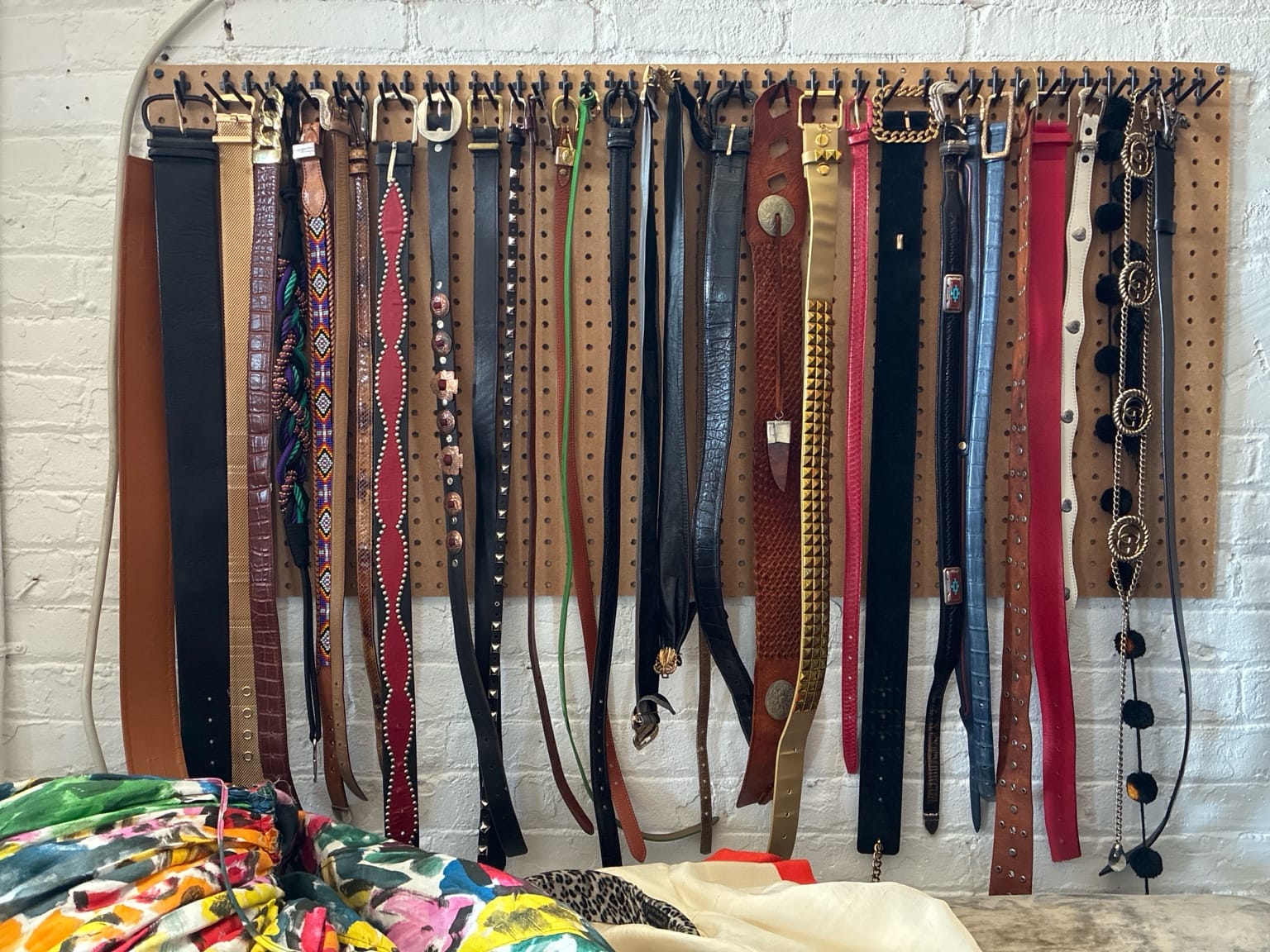A pegboard mounted on a white brick wall, neatly lined with over two dozen belts in various colors, materials, and styles—leather, chain, studded, embroidered. Below the belts, a stack of vibrant, patterned garments rests in the foreground. The belts appear carefully arranged, evoking a sense of both archive and altar.