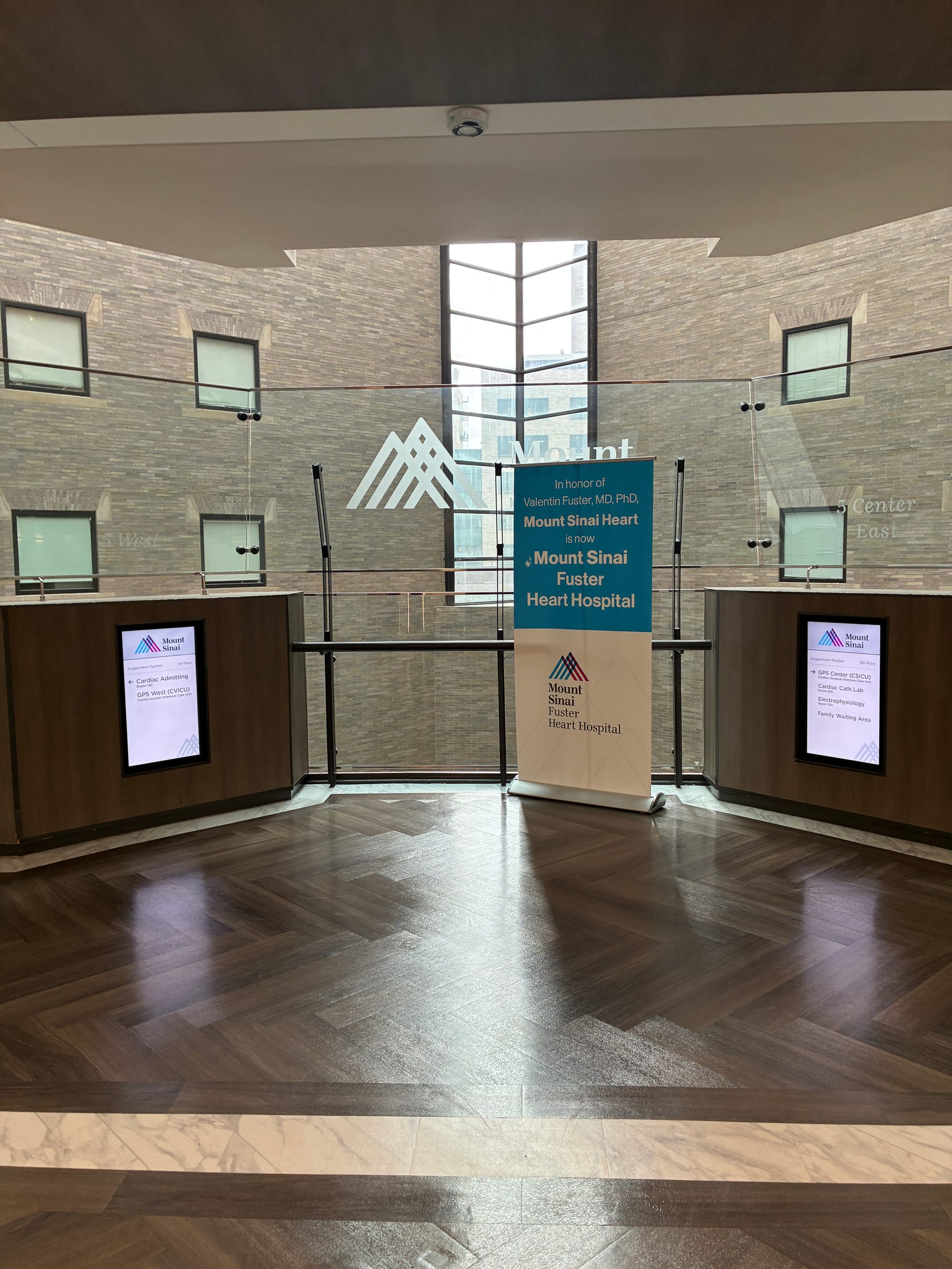 Interior hospital atrium with dark wood floors, Mount Sinai signage, and a banner reading “Mount Sinai Fuster Heart Hospital,” taken on the ICU floor.