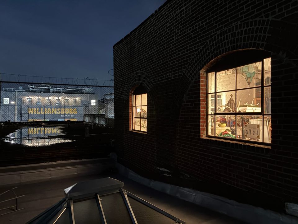 Brooklyn rooftop at night. A lit studio glows through arched windows. Across the fence, “WILLIAMSBURG EATS” is painted on a warehouse wall under a deep blue sky.