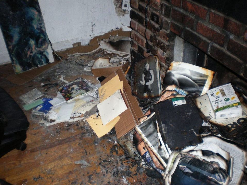 burned apartment floor with scattered papers, books, and cardboard near a damaged brick wall