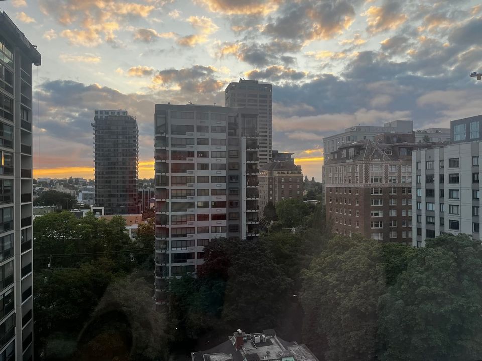 View of a city skyline at dawn, with high-rise apartment buildings in the foreground, trees below, and a partly cloudy sky glowing with orange and pink light at the horizon.