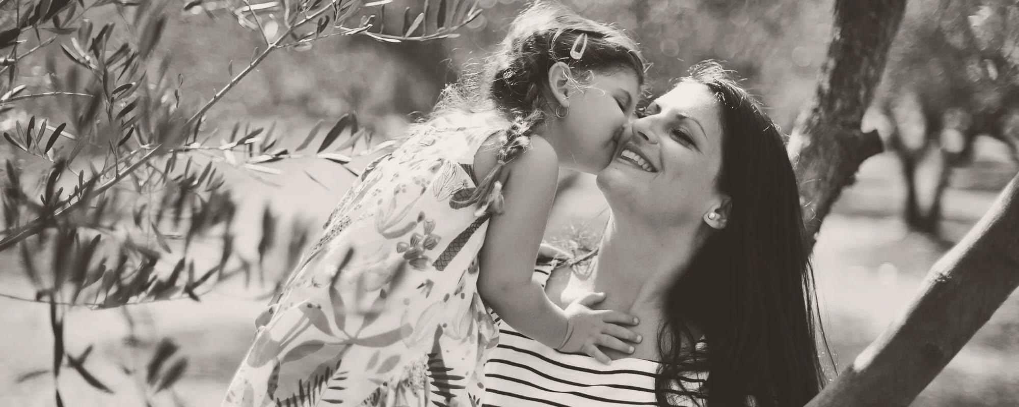 A woman and a young girl sharing a kiss outdoors near a tree, with leaves blurred in the foreground.