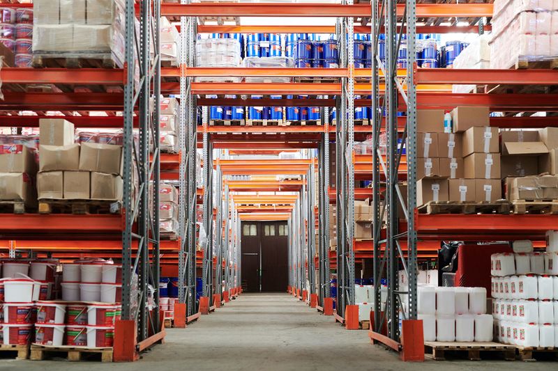 Warehouse aisle with tall orange metal racks stocked with boxed and palletized items.