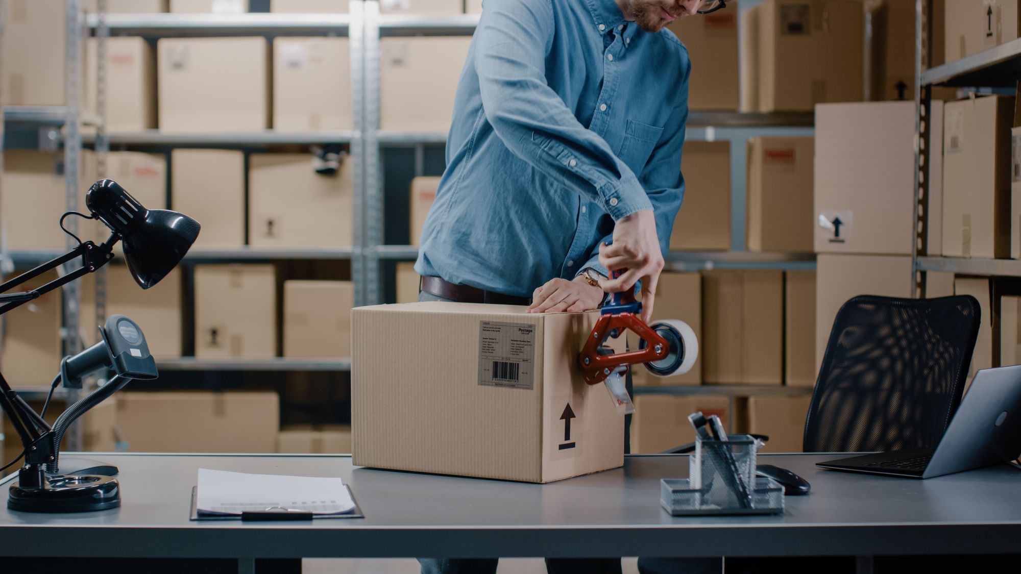 Warehouse worker sealing a cardboard box with tape dispenser for shipping and inventory fulfillment
