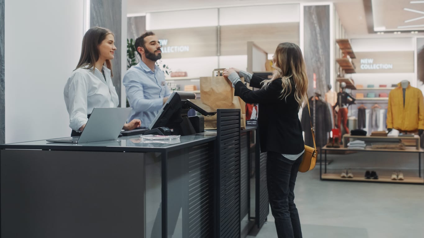Clothing Store Checkout Cashier Counter Young Woman Returns Item in a shopping bag to a Friendly Retail Sales assistant